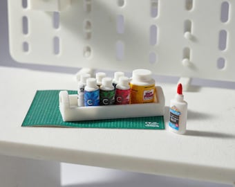 May include: Miniature craft supplies including small bottles of paint in various colors, a bottle of glue with a red tip, and a container of yellow paint. The items are arranged on a green cutting mat and a white tray.