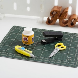 May include: A miniature craft scene with a green cutting mat, a yellow utility knife, a bottle of glue, a stapler, and yellow scissors. A wooden "Home" sign is in the background.