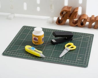 May include: A miniature craft scene with a green cutting mat, a yellow utility knife, a bottle of glue, a stapler, and yellow scissors. A wooden "Home" sign is in the background.