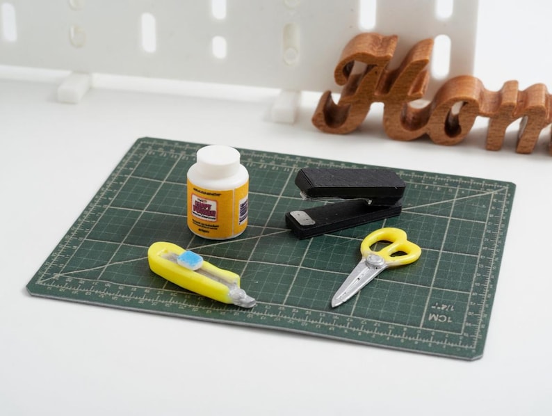May include: A miniature craft scene with a green cutting mat, a yellow utility knife, a bottle of glue, a stapler, and yellow scissors. A wooden "Home" sign is in the background.