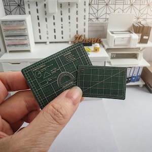 May include: Miniature green cutting mats with grid lines and geometric shapes. One mat has a protractor design. The mats are held in a hand, suggesting a crafting or hobby theme. The background shows a miniature office setting.