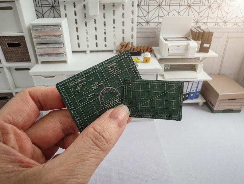May include: Miniature green cutting mats with grid lines and geometric shapes. One mat has a protractor design. The mats are held in a hand, suggesting a crafting or hobby theme. The background shows a miniature office setting.