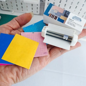 May include: A hand holds a white Easycut paper cutting machine and several colored cutting mats. The mats are pink, blue, and yellow. The machine has a label that reads "Easycut Paper cutting machine".