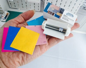 May include: A hand holds a white Easycut paper cutting machine and several colored cutting mats. The mats are pink, blue, and yellow. The machine has a label that reads "Easycut Paper cutting machine".