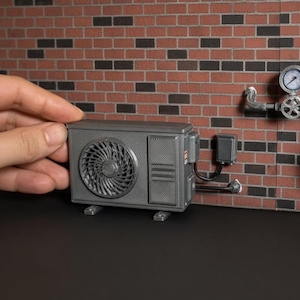 May include: A miniature grey air conditioning unit featuring a circular fan grille and rectangular side vents. The unit is connected to a small electrical box with black wiring and is positioned near a pipe assembly with a round pressure gauge.