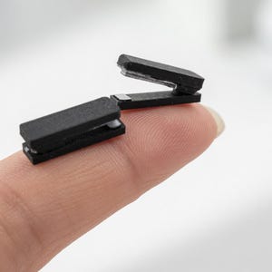 May include: Two small, black staplers are shown. One is open, and the other is closed. The staplers are resting on a fingertip, with a blurred white background.