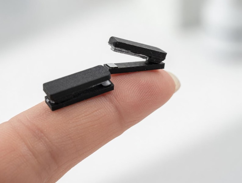 May include: Two small, black staplers are shown. One is open, and the other is closed. The staplers are resting on a fingertip, with a blurred white background.