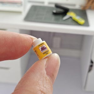 May include: A miniature yellow and white container labeled "Spicy Squid" is held between fingers. The container has a yellow background with black and red text. The background includes a small graphic. The container is in front of a white desk.