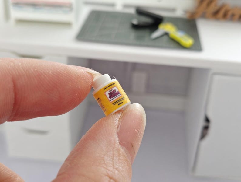 May include: A miniature yellow and white container labeled "Spicy Squid" is held between fingers. The container has a yellow background with black and red text. The background includes a small graphic. The container is in front of a white desk.