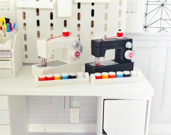 May include: Miniature sewing machines, one white and one black, sit on a white table. Each machine has a red spool of thread and a tray of colorful thread spools. The machines have a silver needle and a white "W" on the side.