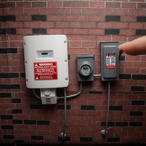 May include: A miniature white solar inverter with a red warning label, a small grey electric meter, and a grey disconnect switch mounted on a brick wall. The red warning label reads WARNING ELECTRICAL SHOCK HAZARD. The components are connected by grey conduits.