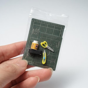 May include: Miniature craft supplies in a clear plastic bag. Includes a green cutting mat, yellow scissors, a yellow utility knife, and a small bottle of glue. The items are designed to be used for crafting or model making.