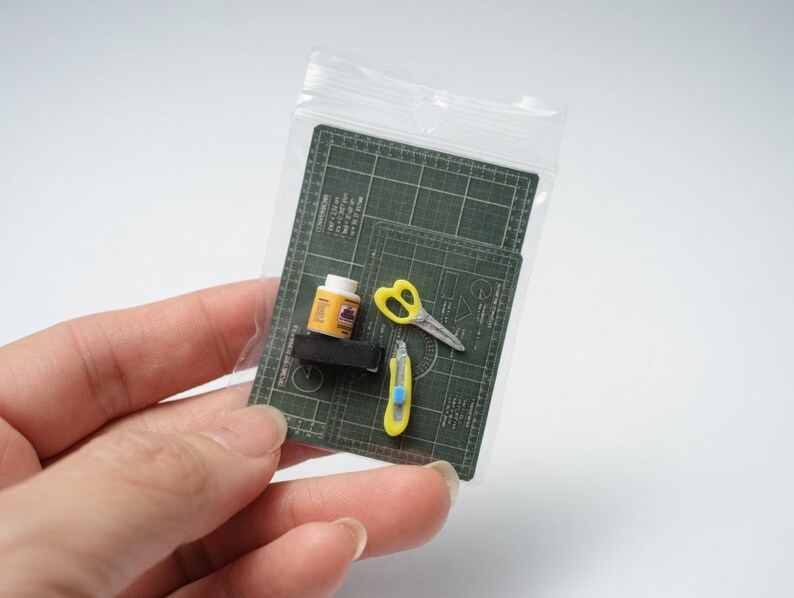 May include: Miniature craft supplies in a clear plastic bag. Includes a green cutting mat, yellow scissors, a yellow utility knife, and a small bottle of glue. The items are designed to be used for crafting or model making.