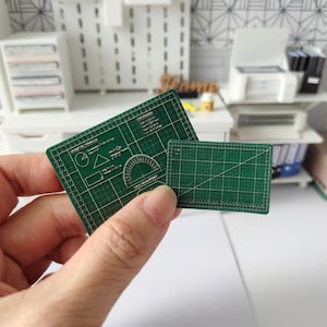 May include: Two miniature green cutting mats are held in a hand. The larger mat has geometric shapes and a protractor printed on it. The smaller mat has a grid pattern. The mats are for a miniature craft or hobby project.
