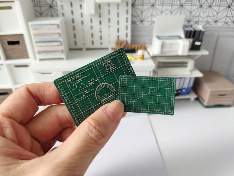 May include: Two miniature green cutting mats are held in a hand. The larger mat has geometric shapes and a protractor printed on it. The smaller mat has a grid pattern. The mats are for a miniature craft or hobby project.