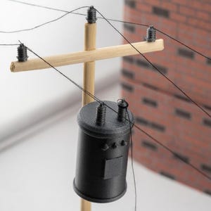May include: A miniature wooden utility pole featuring a horizontal crossarm with three black insulators and thin black wires attached. A cylindrical black transformer is mounted to the pole, also featuring two black insulators on its top surface.
