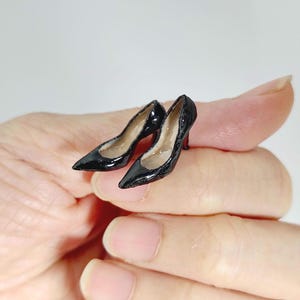 May include: A pair of miniature black high-heeled pumps with a glossy finish, pointed toes, and a distinct red sole detail. These tiny shoes are crafted with a smooth, reflective surface and feature a classic stiletto silhouette in a small scale.