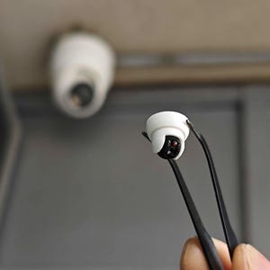 May include: A miniature white and black security camera replica held by black tweezers. The tiny device features a rounded white housing with a black lens section containing a small red dot, mirroring the larger white security camera visible in the background.