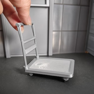May include: A miniature grey plastic flatbed cart with a tall handle and a rectangular platform base. The cart features a textured surface on the platform and a mesh-like design on the handle frame, designed for small-scale display or hobby use.