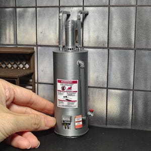May include: A miniature silver water heater model featuring a cylindrical tank, vertical piping, and a red-bordered warning label with text. The unit includes a small control box at the base and a red valve handle on the side, replicating a standard appliance.