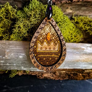 May include: Wooden teardrop pendant with a crown design. The pendant is intricately carved with geometric patterns and a gold-colored crown. It hangs from a black cord with a dark purple bead. The background features moss and wood.