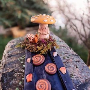 May include: A miniature mushroom with white spots and a brown stem sits on a bed of moss. A small snail figurine is nestled next to the mushroom. A dark blue path with brown swirl decorations leads away from the mushroom.
