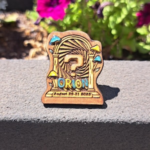 May include: Wooden pin with a carved design featuring the word "ORION" in teal, a question mark, and stylized mushrooms. The pin also includes the text "August 29-31 2025". The pin is set against a grey surface with pink flowers in the background.