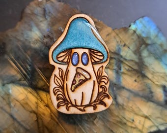 Puede incluir: Pin en forma de hongo de madera con una tapa de color verde azulado y una porción de pizza. El hongo tiene un color de madera clara con contornos y detalles en marrón oscuro. El fondo es una superficie de piedra.