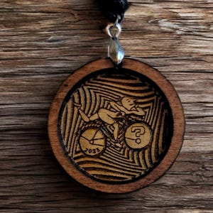 May include: A wooden pendant necklace with a black cord. The pendant is round and features a laser-engraved design of a person riding a bicycle with the year 2023 and a question mark.