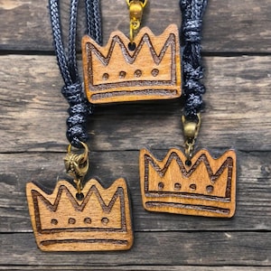 May include: Three wooden crown pendants with black cords and gold-colored clasps. The crowns are carved with a detailed design and have three small circular accents. The pendants are displayed on a weathered wooden surface.