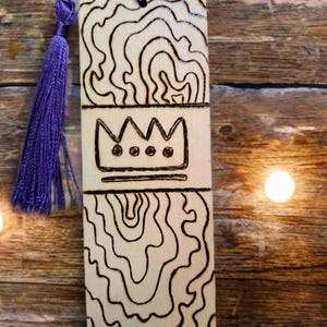 May include: A wooden bookmark with a purple tassel. The bookmark has a wood-burned design of a crown with three jewels and a wavy pattern.