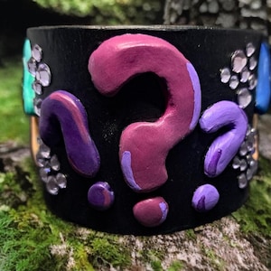 Op de afbeelding: Zwarte leren armband met paarse, roze en blauwe vraagtekens en strass.