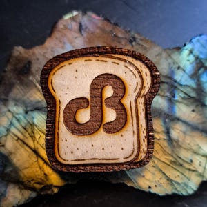 May include: A wooden bread-shaped pin with a toasted brown border and a light tan center. The center features a stylized brown letter "dB" design. The pin is set against a colorful, textured background.