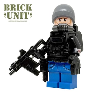 May include: A minifigure wearing a gray beanie, a black tactical vest, and blue pants. The figure has a black respirator mask, a black rifle, and a black backpack. The text BRICK UNIT CUSTOM ACTION MINIFIGURES is printed in the upper left corner.