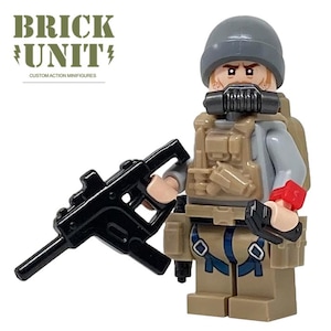 May include: A custom action minifigure of a soldier with a gray helmet, gas mask, and tan tactical vest. The minifigure holds a black submachine gun and a black sidearm. The text "BRICK UNIT" and "CUSTOM ACTION MINIFIGURES" are visible.