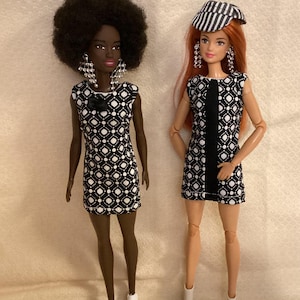 May include: Two fashion dolls wearing black and white geometric print dresses. One doll has long red hair and is wearing a black and white striped hat. The other doll has short black curly hair. Both dolls are wearing white shoes.