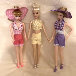 May include: Three Barbie dolls wearing summer outfits. The doll on the left is wearing a pink and white gingham halter top, pink shorts, and a pink and white polka dot hat. The doll in the middle is wearing a yellow and gold patterned crop top, yellow shorts, and a white headband. The doll on the right is wearing a purple and white gingham halter top, purple shorts, and a white and purple polka dot hat.