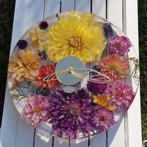 Peut inclure: Une horloge ronde avec un revêtement en résine transparent, ornée de fleurs pressées jaunes, oranges et violettes. Le cadran est argenté et les aiguilles dorées. Diamètre d'environ 25 cm.