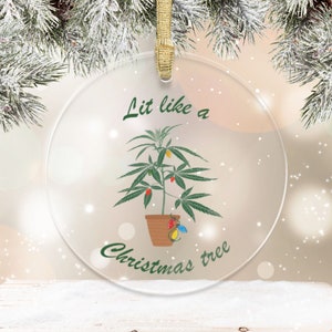 May include: Clear glass Christmas bauble featuring a cannabis plant design. The plant is adorned with festive lights, and the text "Lit like a Christmas tree" is written in green. A gold ribbon is attached for hanging.