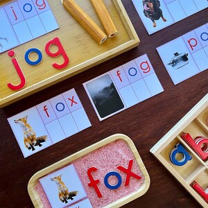 Pode incluir: Um conjunto de flashcards educacionais com imagens e palavras para as palavras "jog", "fox", "fog" e "pot". Os cart&otilde;es est&atilde;o dispostos em uma mesa de madeira com uma bandeja de pe&ccedil;as de letras e uma bandeja de madeira com pe&ccedil;as de letras vermelhas e azuis que soletram a palavra "fox".