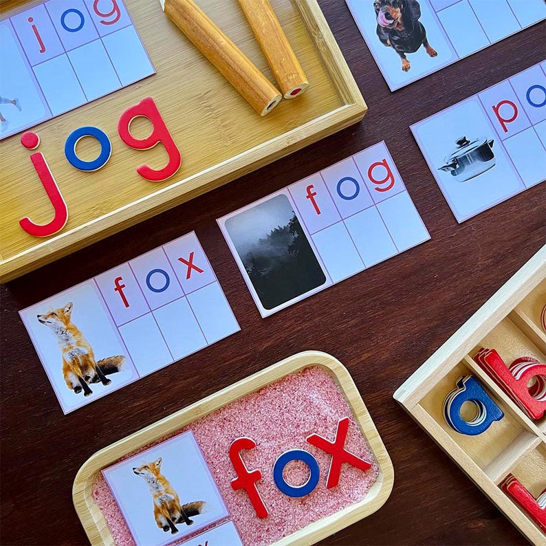 Pode incluir: Um conjunto de flashcards educacionais com imagens e palavras para as palavras "jog", "fox", "fog" e "pot". Os cart&otilde;es est&atilde;o dispostos em uma mesa de madeira com uma bandeja de pe&ccedil;as de letras e uma bandeja de madeira com pe&ccedil;as de letras vermelhas e azuis que soletram a palavra "fox".