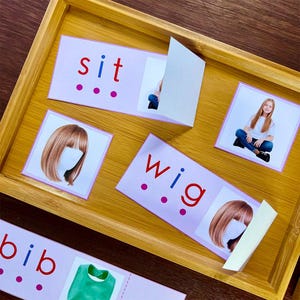 Pode incluir: Um conjunto de flashcards rosa e branco com as palavras "sit", "wig" e "bib" impressas nelas. Cada cart&atilde;o tem uma imagem correspondente, como uma pessoa sentada, uma peruca e um babador.