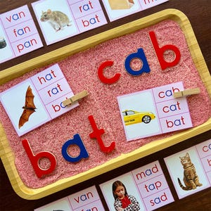 Pode incluir: Uma bandeja de madeira com areia rosa e letras magn&eacute;ticas coloridas que soletram as palavras "bat" e "cab". Existem v&aacute;rios cart&otilde;es pequenos com palavras e imagens neles, incluindo um morcego, um carro, um gato e um chap&eacute;u.