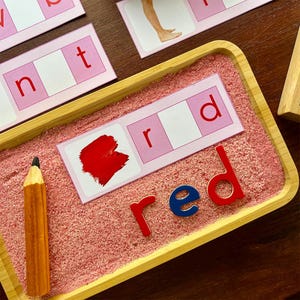 Pode incluir: Um cart&atilde;o rosa e branco com a palavra "red" escrita nele. As letras est&atilde;o em uma amostra de tinta vermelha, uma letra vermelha "r", um espa&ccedil;o em branco e uma letra vermelha "d". A palavra "red" est&aacute; escrita com letras magn&eacute;ticas em uma bandeja de madeira cheia de areia vermelha.
