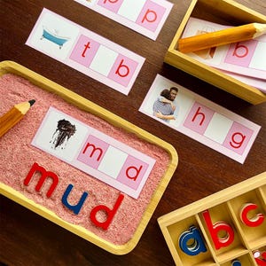 Pode incluir: Um cart&atilde;o rosa e branco com a palavra "mud" escrita em letras vermelhas e azuis. O cart&atilde;o est&aacute; em uma bandeja com areia rosa. H&aacute; uma imagem de lama no cart&atilde;o.