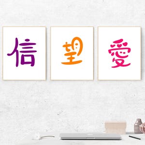 May include: Three framed prints with Chinese characters in purple, orange, and pink. The characters are displayed on a white background with a light wooden frame. The prints are arranged on a white wall above a desk with a laptop and decorative items.