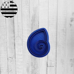 Può includere: Tagliabiscotti blu a forma di conchiglia con un motivo a spirale. Il tagliabiscotti ha un bordo esterno arrotondato. Lo sfondo è una superficie di legno di colore chiaro. Un logo in bianco e nero si trova nell'angolo in alto a sinistra.