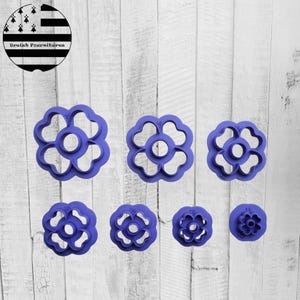 Peut inclure: Ensemble de sept emporte-pièces en forme de fleur de couleur violette, de différentes tailles. Les emporte-pièces ont un trou central et des découpes en forme de pétales. Un logo circulaire avec du texte et un design noir et blanc est également présent.