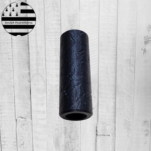 Op de afbeelding: Een zwart cilindrisch object met een textuuroppervlak. Het object staat rechtop tegen een witte houten achtergrond. Het object lijkt een decoratief item of een onderdeel van een groter product te zijn.