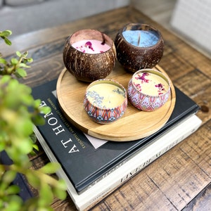 Peut inclure: Quatre bougies de diff&eacute;rentes couleurs et designs, dont une bougie bleue dans une coque de noix de coco, une bougie rose dans une coque de noix de coco, une bougie jaune dans une bo&icirc;te &agrave; motifs et une bougie jaune dans une bo&icirc;te &agrave; motifs. Les bougies sont dispos&eacute;es sur un plateau en bois avec un livre en dessous.